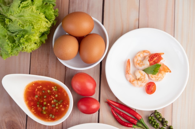 Drei frische Garnelen, Eier, Chili, Sauce und halbe Tomaten in einem weißen Teller auf einem Holz.