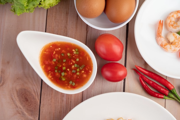 Drei frische Garnelen, Eier, Chili, Sauce und halbe Tomaten in einem weißen Teller auf einem Holz.