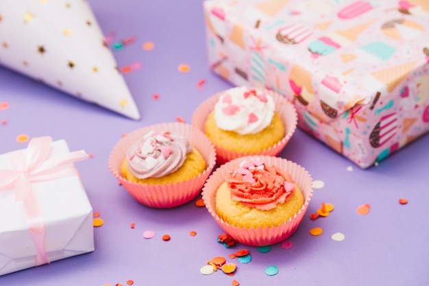 Kostenloses Foto drei gebackene muffins mit geschenken; partyhut und konfetti auf lila hintergrund