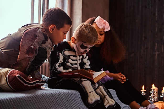 Drei gemischtrassige Kinder in gruseligen Kostümen lesen Horrorgeschichten, während sie in einem alten Haus auf dem Bett sitzen. Halloween-Konzept.