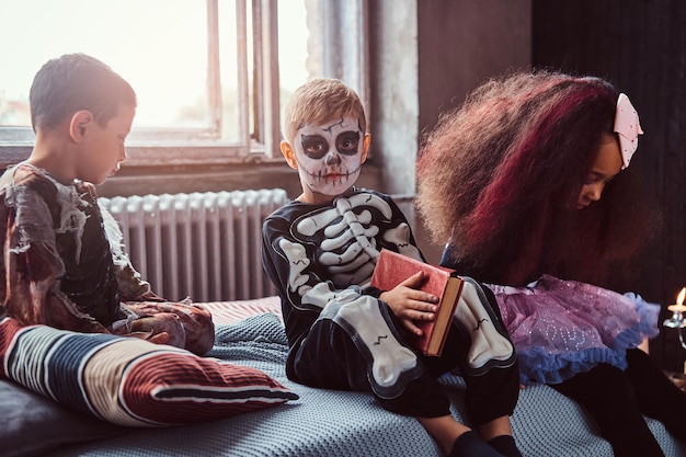 Drei gemischtrassige Kinder in gruseligen Kostümen lesen Horrorgeschichten, während sie in einem alten Haus auf dem Bett sitzen. Halloween-Konzept.
