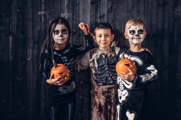 Drei gemischtrassige Kinder in gruseligen Kostümen posieren mit Kürbissen in einem alten Haus. Halloween-Konzept.