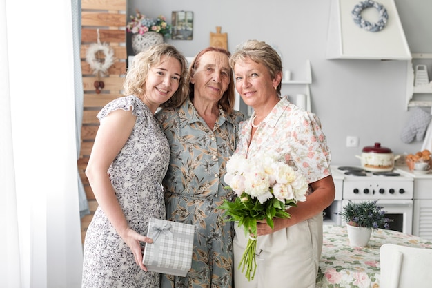 Drei Generationsfrauen, die zusammen stehen, Blumenblumenstrauß und -geschenk halten, die Kamera betrachten