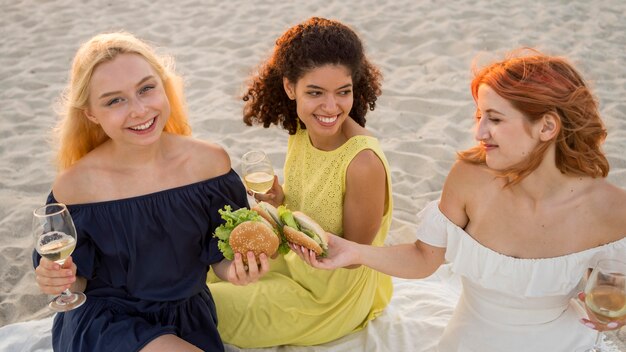 Drei glückliche Freundinnen, die zusammen Burger am Strand genießen