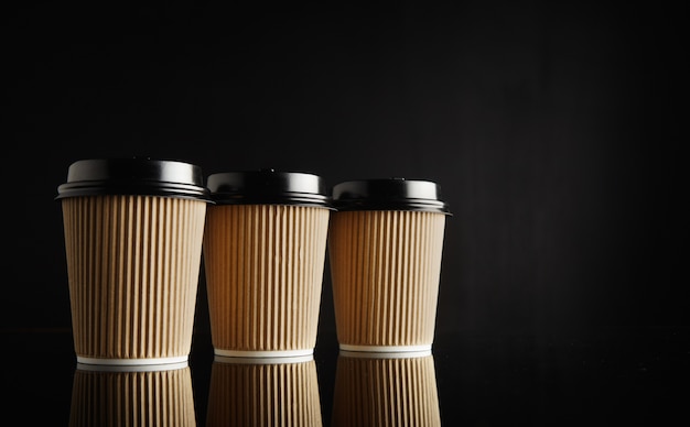 Drei identische hellbraune Pappkaffeetassen zum Mitnehmen mit schwarzen Deckeln in einer Reihe auf einem reflektierenden schwarzen Tisch gegen eine schwarze Wand