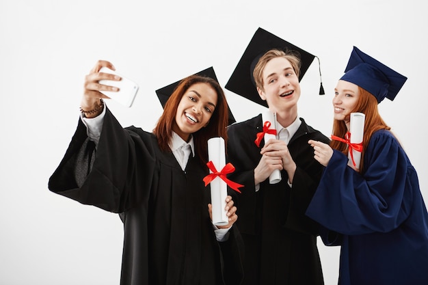 Drei internationale Absolventenfreunde freuen sich über Mäntel und machen ein Selfie am Telefon. Zukünftige Spezialisten haben Spaß mit ihren Diplomen.