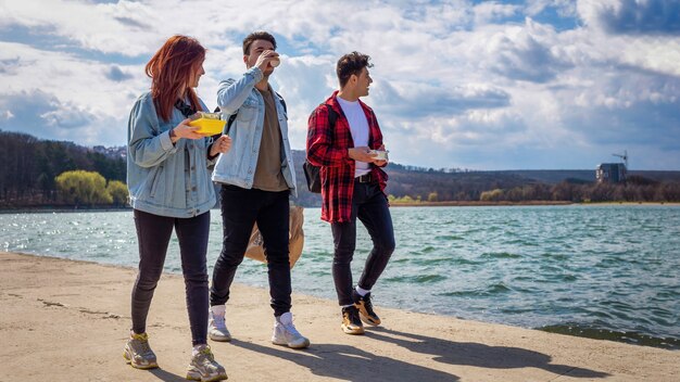 Drei junge Freunde gehen am See entlang, trinken und essen in einem Park