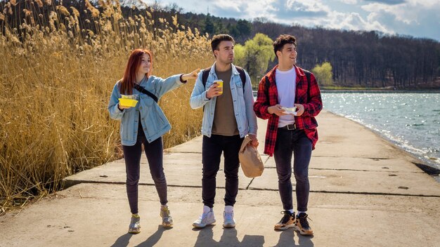 Drei junge Freunde gehen am See entlang, trinken und essen in einem Park