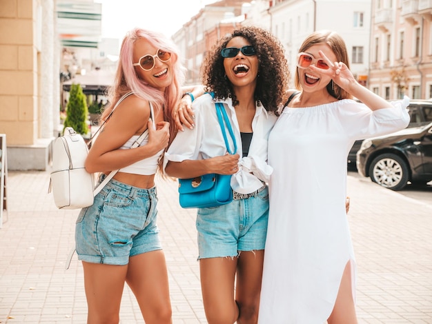 Drei junge schöne lächelnde Frauen in trendiger SommerkleidungSexy sorglose gemischtrassige Frauen posieren auf der Straße HintergrundPositive Models, die Spaß mit Sonnenbrillen haben Fröhlich und glücklich