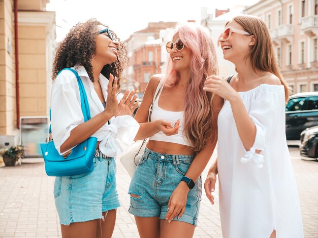 Drei junge schöne lächelnde Frauen in trendiger SommerkleidungSexy sorglose gemischtrassige Frauen posieren auf der Straße HintergrundPositive Models, die Spaß mit Sonnenbrillen haben Fröhlich und glücklich