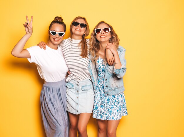 Drei junge schöne lächelnde Hippie-Mädchen in der modischen Sommerkleidung. Sexy sorglose Frauen, die nahe gelber Wand aufwerfen. Positive Modelle, die Spaß haben. Sie zeigen Friedenszeichen