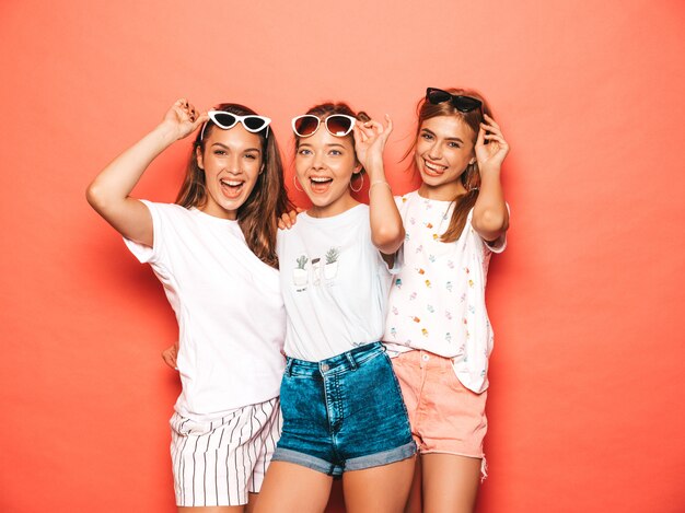 Drei junge schöne lächelnde Hippie-Mädchen in der modischen Sommerkleidung. Sexy sorglose Frauen, die nahe rosa Wand aufwerfen. Positive Models werden verrückt und haben Spaß mit Sonnenbrillen