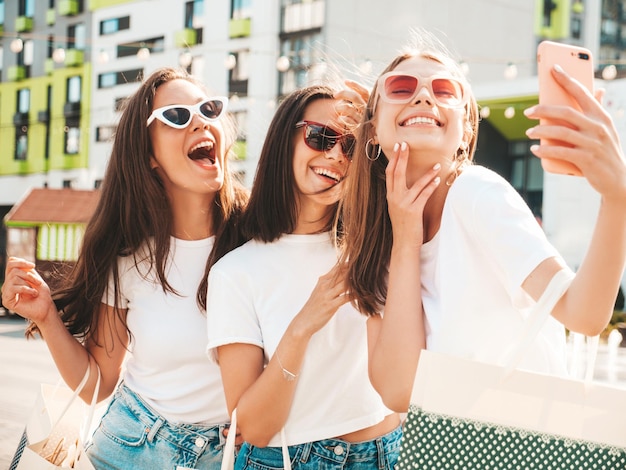 Drei junge schöne lächelnde Hipster-Frauen im trendigen Sommer gleiche Kleidung Sexy sorglose Frauen posieren auf der StraßePositive Models, die Spaß mit Sonnenbrillen habenEinkaufstaschen haltenSelfie-Fotos machen