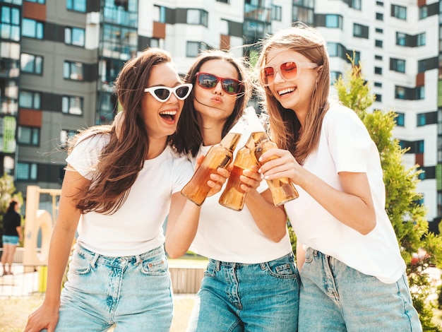 Drei junge schöne lächelnde Hipster-Frauen im trendigen Sommer gleiche KleidungSexy unbeschwerte Frauen posieren auf der StraßePositive Models, die Spaß mit Sonnenbrillen haben Trinkflasche Bier Oktoberfest