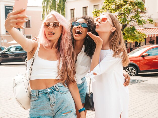 Drei junge schöne lächelnde Hipster-Frauen in trendiger SommerkleidungSexy sorglose gemischtrassige Frauen posieren auf dem StraßenhintergrundPositive Models, die Spaß mit Sonnenbrillen haben Selfie-Fotos machen