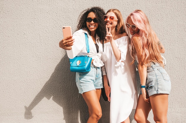 Drei junge schöne lächelnde Hipster-Frauen in trendiger SommerkleidungSexy sorglose gemischtrassige Frauen posieren auf dem StraßenhintergrundPositive Models, die Spaß mit Sonnenbrillen haben Selfie-Fotos machen