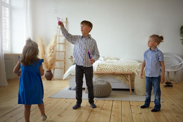 Drei Kinder spielen zu Hause zusammen. Schuljunge in Hemd und Jeans bläst Seifenblasen im geräumigen Schlafzimmer, sein kleiner Bruder und seine kleine Schwester warten darauf, dass sie an die Reihe kommen, und stehen um ihn herum auf dem Boden