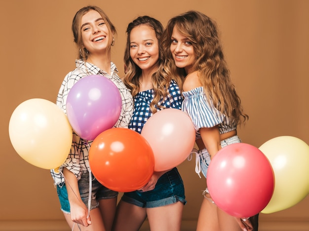 Drei lächelnde Schönheiten im karierten Hemdsommer kleidet. Mädchen posieren. Modelle mit bunten Luftballons. Spaß haben, bereit zum Feiergeburtstag