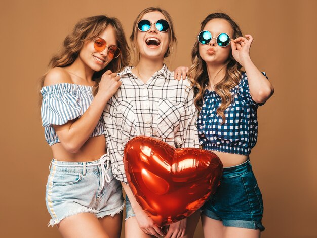 Drei lächelnde Schönheiten im karierten Hemdsommer kleidet. Mädchen posieren. Modelle mit Herzformballon in der Sonnenbrille. Bereiten Sie für Feier Valentinstag vor