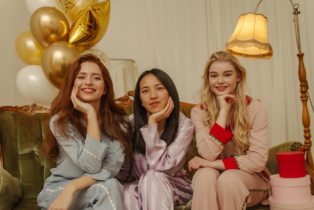Drei schöne junge interrassische Damen tragen farbenfrohe Pyjamas und schauen in die Kamera, während sie in einem dekorierten Raum sitzen Stilvolles Urlaubskonzept