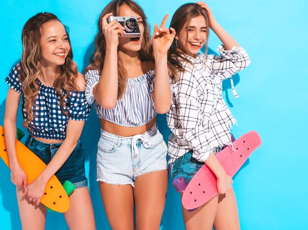 Drei sexy schöne stilvolle lächelnde Mädchen mit bunten Pennyskateboards. Frauen in der karierten Hemdkleidungsaufstellung des Sommers. Modelle, die Fotos auf Retro- Fotokamera machen