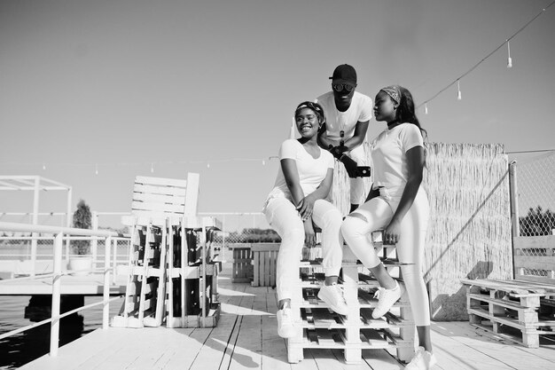Drei stilvolle afroamerikanische Freunde tragen weiße Kleidung am Pier am Strand Straßenmode junger schwarzer Menschen Schwarzer Mann mit zwei afrikanischen Mädchen