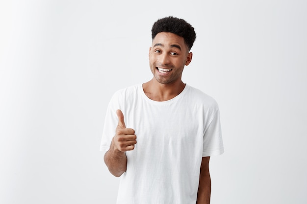 Du siehst gut aus, Mann. Schließen Sie herauf Porträt des reifen gutaussehenden dunkelhäutigen fröhlichen Mannes mit Afro-Frisur im lässigen weißen T-Shirt, das mit Zähnen lächelt und Daumen hoch Zeichen mit glücklichem Ausdruck zeigt.