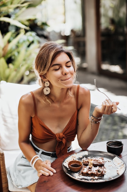 Dünn gebräunte Frau in braunem BH und stylischen Jeansshorts schmeckt nach Waffel mit Sahne, Erdnüssen und Ahornsirup