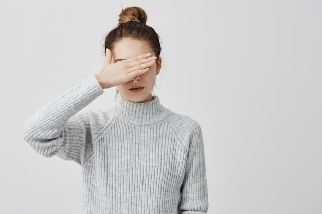 Dünne Frau, die graues Outfit trägt und ihre Augen mit der Hand schließt. Selbstbewusste Frauen, die versuchen, ihr Gesicht vor anderen Menschen zu verbergen, ziehen es vor, nicht zu sehen. Entscheidung, menschliches Konzept