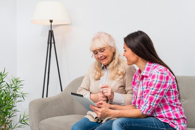Dumme ältere Mutter und ihre Tochter, die eine digitale Tablette beim Sitzen auf Sofa verwendet