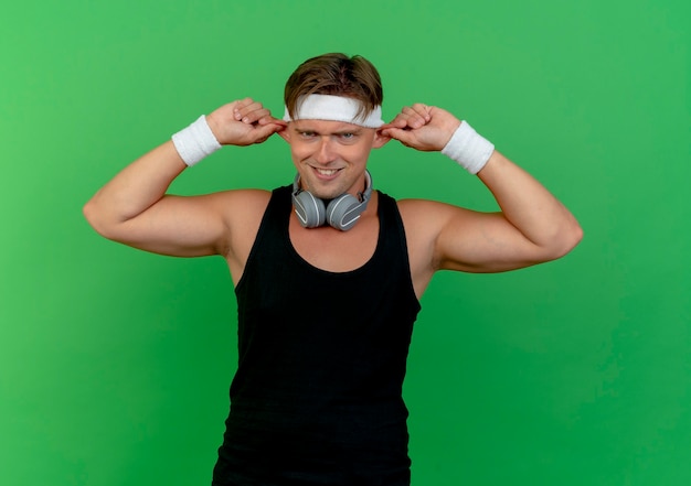Dumme junge hübsche sportliche mann, die stirnband und armbänder mit kopfhörern am hals trägt, die affenohren lokalisiert auf grüner wand machen