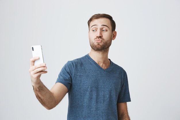 Dummer hübscher Kerl, der Selfie auf Smartphone nimmt und schmollt