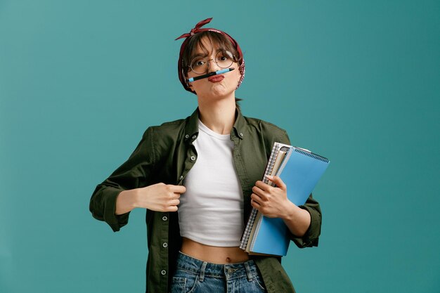 dummes junges studentenmädchen, das eine bandana-brille trägt, die große notizblöcke hält, die ihr hemd greifen und einen schnurrbart mit einem stift machen, der isoliert auf blauem hintergrund in die kamera schaut