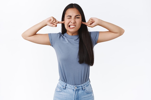 Dummes und kindisches süßes asiatisches Mädchen weigert sich, Ihnen zuzuhören, wenn Sie die Ohren mit den Fingern bedecken, zusammenzucken, schielen und die Zähne zusammenbeißen, wenn Sie stören, störende laute Geräusche, weiße Wand