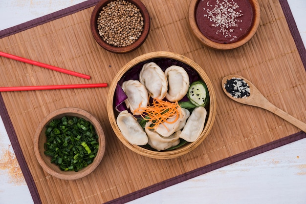 Dumpling mit Salat im Bambusdampfer, umgeben von Schnittlauch; Koriandersamen und Essstäbchen auf Tischset