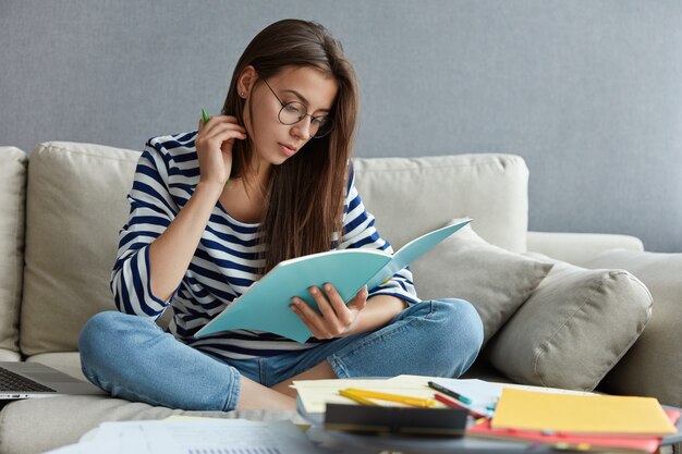 Dunkelhaarige attraktive Frau, die ein Lehrbuch liest, umgeben von Papieren, hält Stift