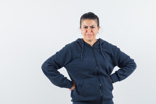 Dunkelhaarige Frau, die Hand auf Taille in Jacke hält und unzufrieden aussieht