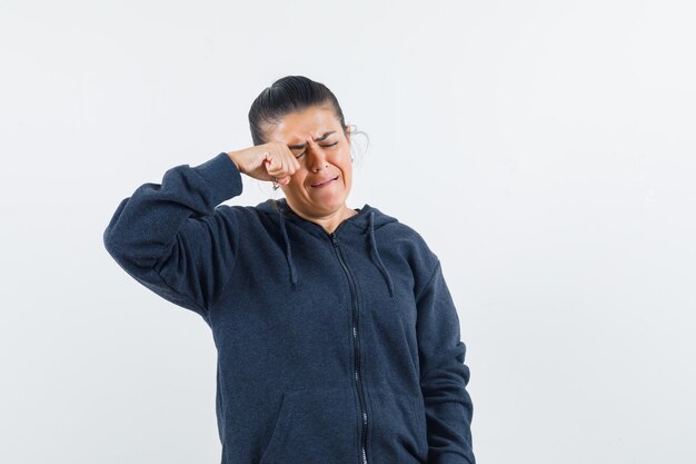 Dunkelhaarige Frau, die sich das Auge reibt, während sie in der Jacke weint und traurig aussieht. Vorderansicht.