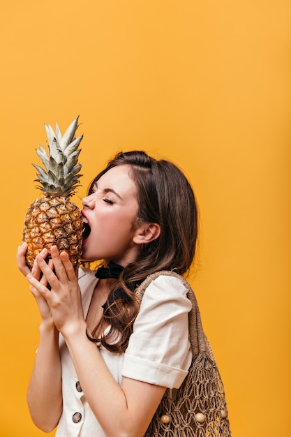 Dunkelhaarige Frau im weißen T-Shirt mit Stringtasche auf der Schulter beißt Ananas.