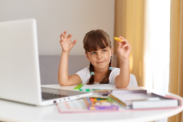 Dunkelhaariges süßes schulmädchen mit weißem t-shirt, das zu hause drinnen posiert, am tisch sitzt, umgeben von büchern, vor laptop-computer, online-bildung.