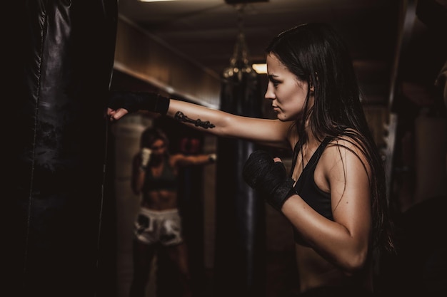 Dunkles Foto einer jungen hübschen Frau in einem dunklen Fitnessstudio, das mit einem Boxsack trainiert.