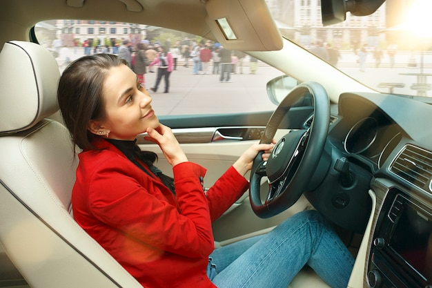 Durch die Stadt fahren. Junge attraktive Frau, die ein Auto fährt. Junges hübsches kaukasisches Modell in der eleganten stilvollen roten Jacke, die am modernen Fahrzeuginnenraum sitzt. Geschäftsfrau Konzept.