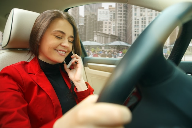 Durch die Stadt fahren. Junge attraktive Frau, die ein Auto fährt. Junges hübsches kaukasisches Modell in der eleganten stilvollen roten Jacke, die am modernen Fahrzeuginnenraum sitzt. Geschäftsfrau Konzept.