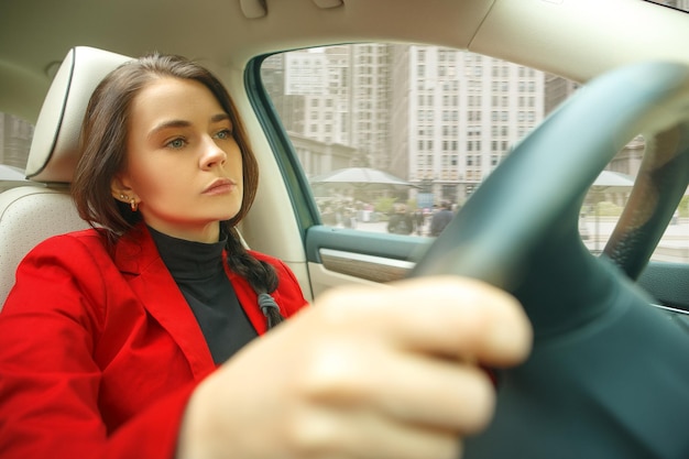 Kostenloses Foto durch die stadt fahren. junge attraktive frau, die ein auto fährt. junges hübsches kaukasisches modell in der eleganten stilvollen roten jacke, die am modernen fahrzeuginnenraum sitzt. geschäftsfrau konzept.