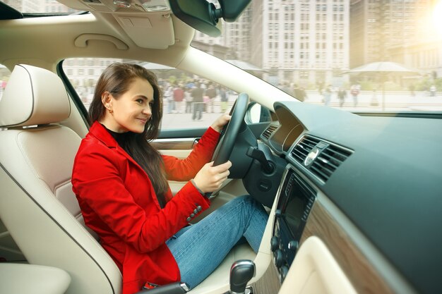 Durch die Stadt fahren. Junge attraktive Frau, die ein Auto fährt. Junges hübsches kaukasisches Modell in der eleganten stilvollen roten Jacke, die am modernen Fahrzeuginnenraum sitzt.