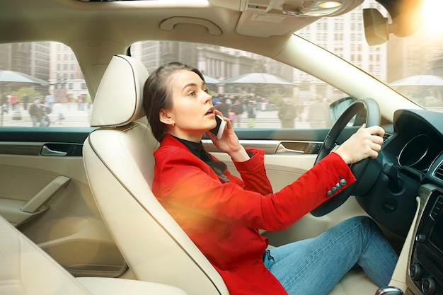 Durch die Stadt fahren. Junge attraktive Frau, die ein Auto fährt. Junges hübsches kaukasisches Modell in der eleganten stilvollen roten Jacke, die am modernen Fahrzeuginnenraum sitzt