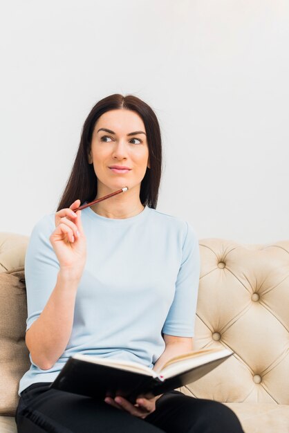 Durchdachte Frau, die mit Notizbuch und Bleistift auf Couch sitzt