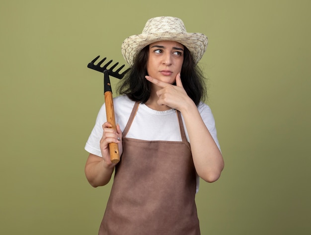 Durchdachte junge brünette Gärtnerin in Uniform, die Gartenhut trägt, legt Hand auf Kinn und hält Rechen, der Seite betrachtet, die auf olivgrüner Wand lokalisiert wird