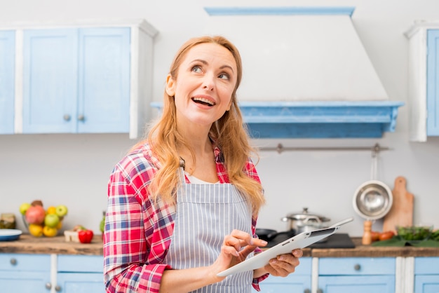 Durchdachte junge Frau, die in der Hand die digitale Tablette steht in der Küche verwendet
