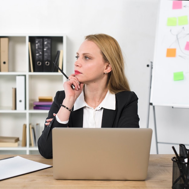 Durchdachte junge Geschäftsfrau, die am Arbeitsplatz mit Laptop auf Tabelle sitzt